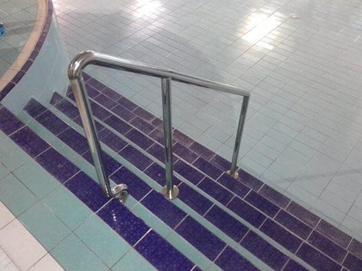 Metalowa poręcz schodów basenowych z niebieskimi i turkusowymi płytkami, widok z góry, oświetlenie sufitowe odbija się na mokrej powierzchni.