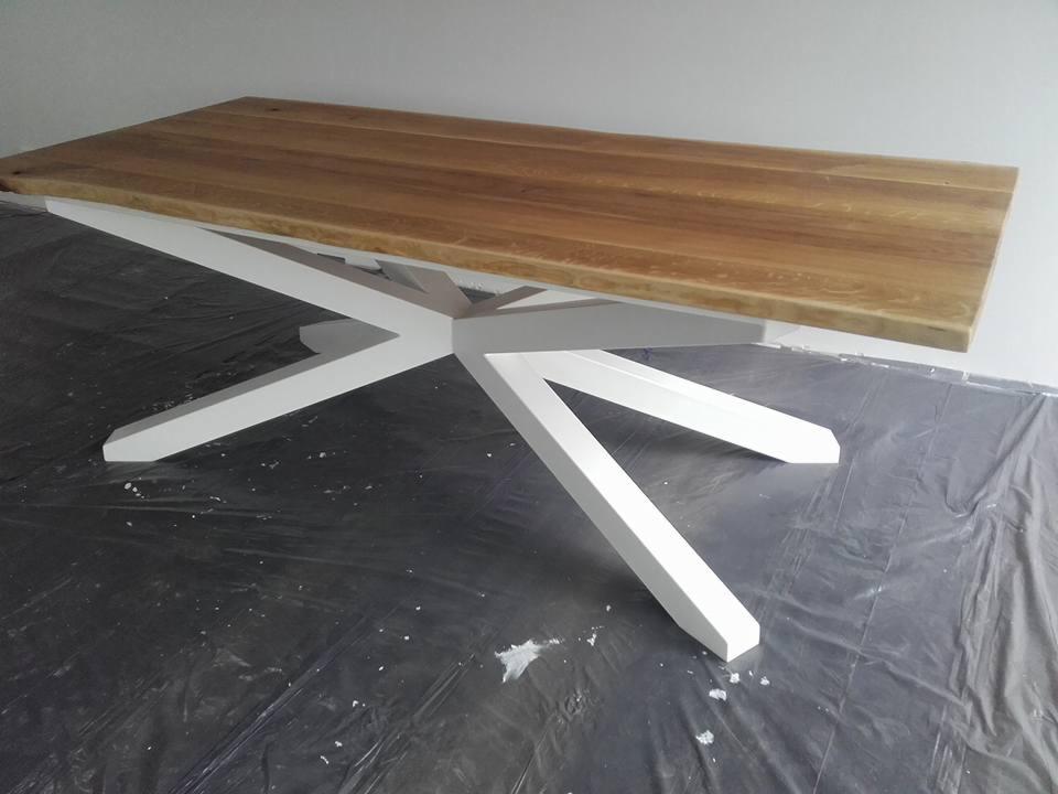 Stół z blatem z jasnego drewna i białą, geometryczną podstawą w kształcie gwiazdy, stojący na folii ochronnej na podłodze. Minimalistyczny design.