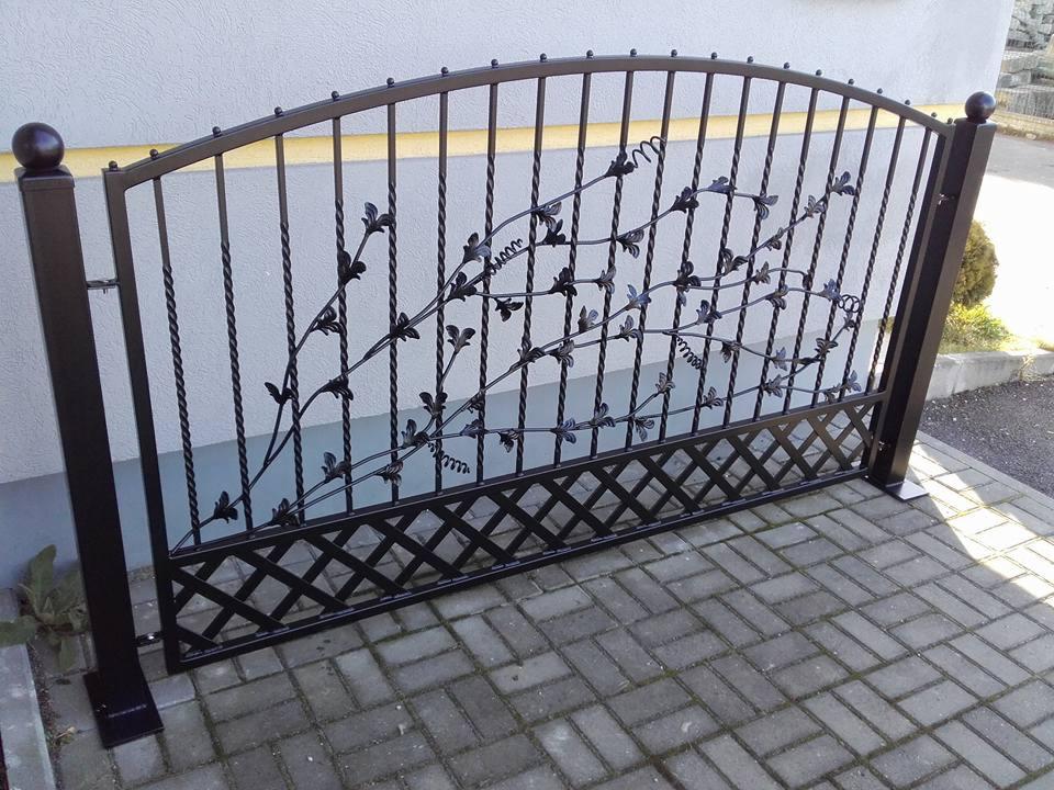 Kuta, czarna brama z motywem roślinnym, widoczna z boku na tle szarej ściany i kostki brukowej. Dekoracyjny wzór z liści na metalowej konstrukcji.