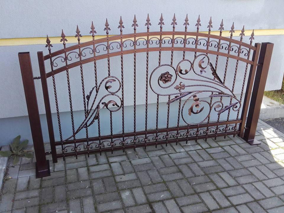 Dekoracyjne, kute przęsło ogrodzeniowe w kolorze brązu, z motywami roślinnymi, ustawione na brukowanej nawierzchni przed jasną ścianą budynku.