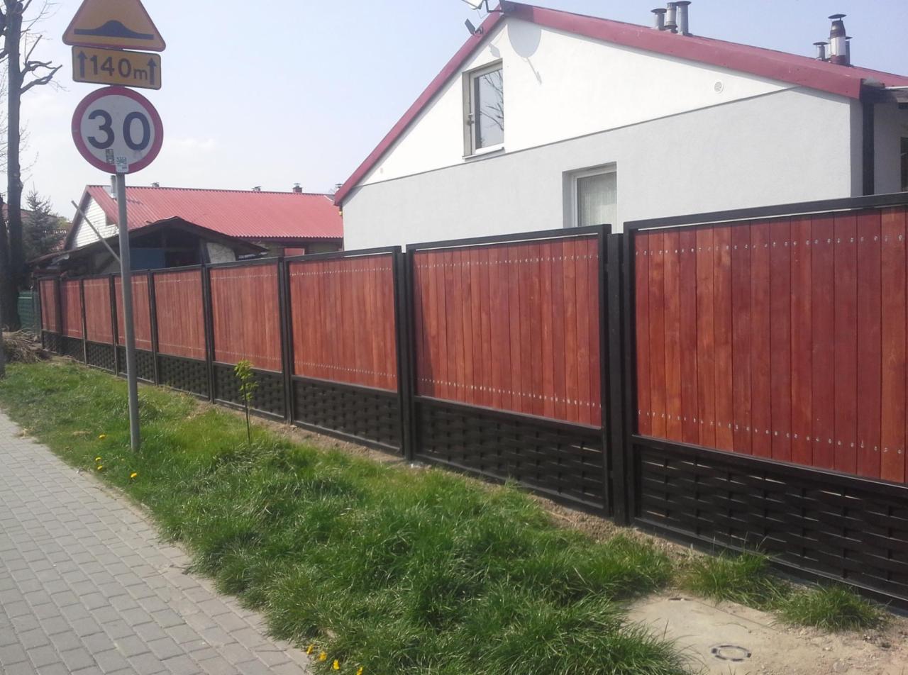 Nowoczesne ogrodzenie panelowe z drewnianym wypełnieniem w odcieniach brązu i czarną, plecioną podstawą, widoczne z poziomu chodnika, z domem w tle.