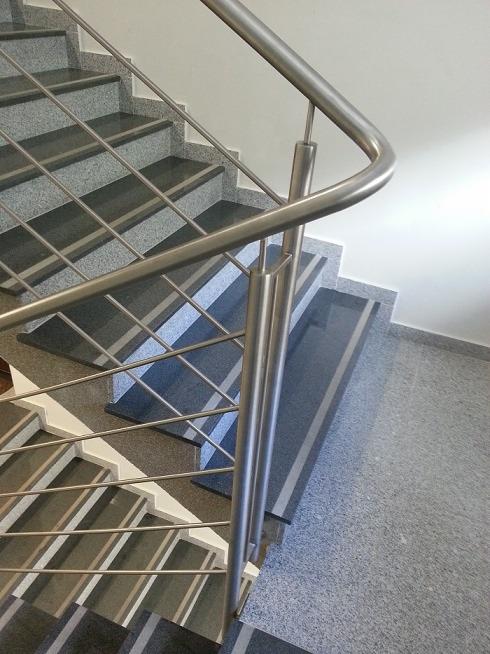 Nowoczesna balustrada nierdzewna ze szczeblami, zamontowana na granitowych schodach wewnątrz budynku. Widok z góry, skupienie na detalu połączeń.