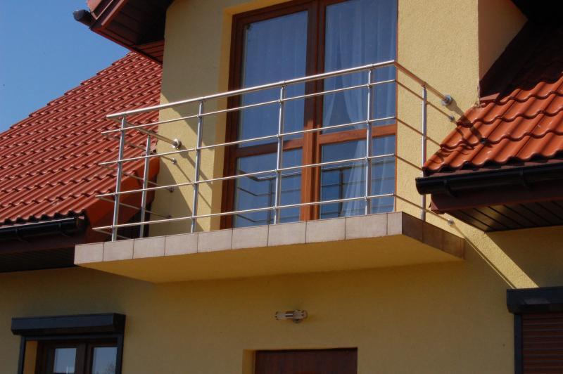 Balkon z nierdzewną balustradą na tle żółtej elewacji domu z czerwoną dachówką, widok z dołu. Balustrada z poziomymi prętami, brązowe ramy okien.