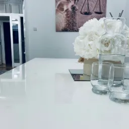 Jasna sala konferencyjna z białym stołem, bukietem białych piwonii w beżowym pudełku, szklanym dzbankiem z wodą i czterema szklankami; na ścianie obraz z motywem Temidy i wagą.