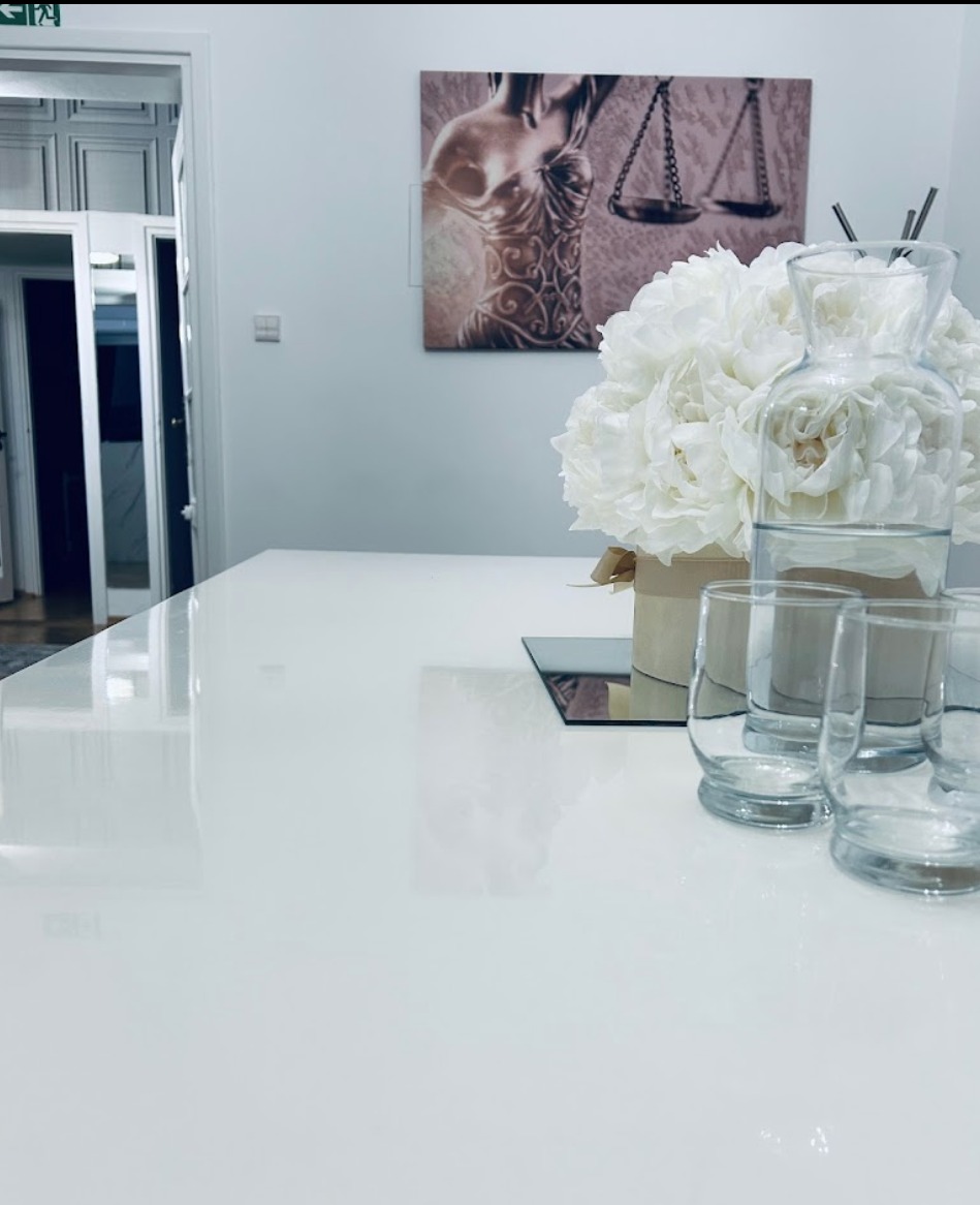 Jasna sala konferencyjna z białym stołem, bukietem białych piwonii w beżowym pudełku, szklanym dzbankiem z wodą i czterema szklankami; na ścianie obraz z motywem Temidy i wagą.