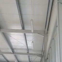 Wnętrze hali produkcyjnej z widocznymi podwieszanymi lampami tubowymi na linkach, stalowa konstrukcja dachu i słupów pomalowana na szaro.