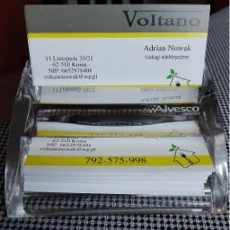 Wizytówki firmy Voltano z Konina, oferującej usługi elektryczne, umieszczone w przezroczystym stojaku na czarno-białym tle. Żółty pasek na wizytówce.
