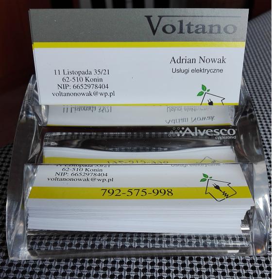 Wizytówki firmy Voltano z Konina, oferującej usługi elektryczne, umieszczone w przezroczystym stojaku na czarno-białym tle. Żółty pasek na wizytówce.