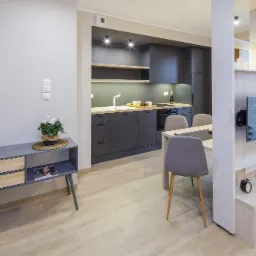 Nowoczesne wnętrze mieszkania z szarą zabudową kuchenną, drewnianym stołem i krzesłami, oraz szafką RTV z telewizorem na ścianie działowej.