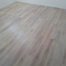 Jasne panele podłogowe ułożone w jodełkę w rogu pokoju, widoczny fragment ściany i gniazdko elektryczne. Nowoczesne wykończenie wnętrza.