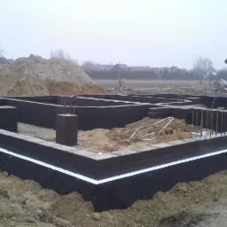 Zabezpieczone fundamenty domu w trakcie budowy, widoczne zbrojenia wystające z betonowych słupków, izolacja pionowa z folii kubełkowej z białym paskiem na górze, w tle hałda ziemi i częściowo...