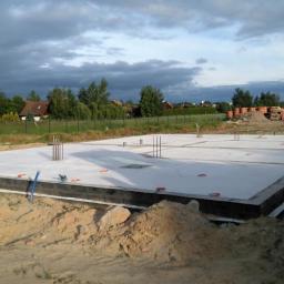 Piotr Jędrzejczak Usługi Ogólnobudowlane - Wylana płyta fundamentowa z wystającymi prętami zbrojeniowymi na działce z widokiem na domy w oddali pod pochmurnym niebem.