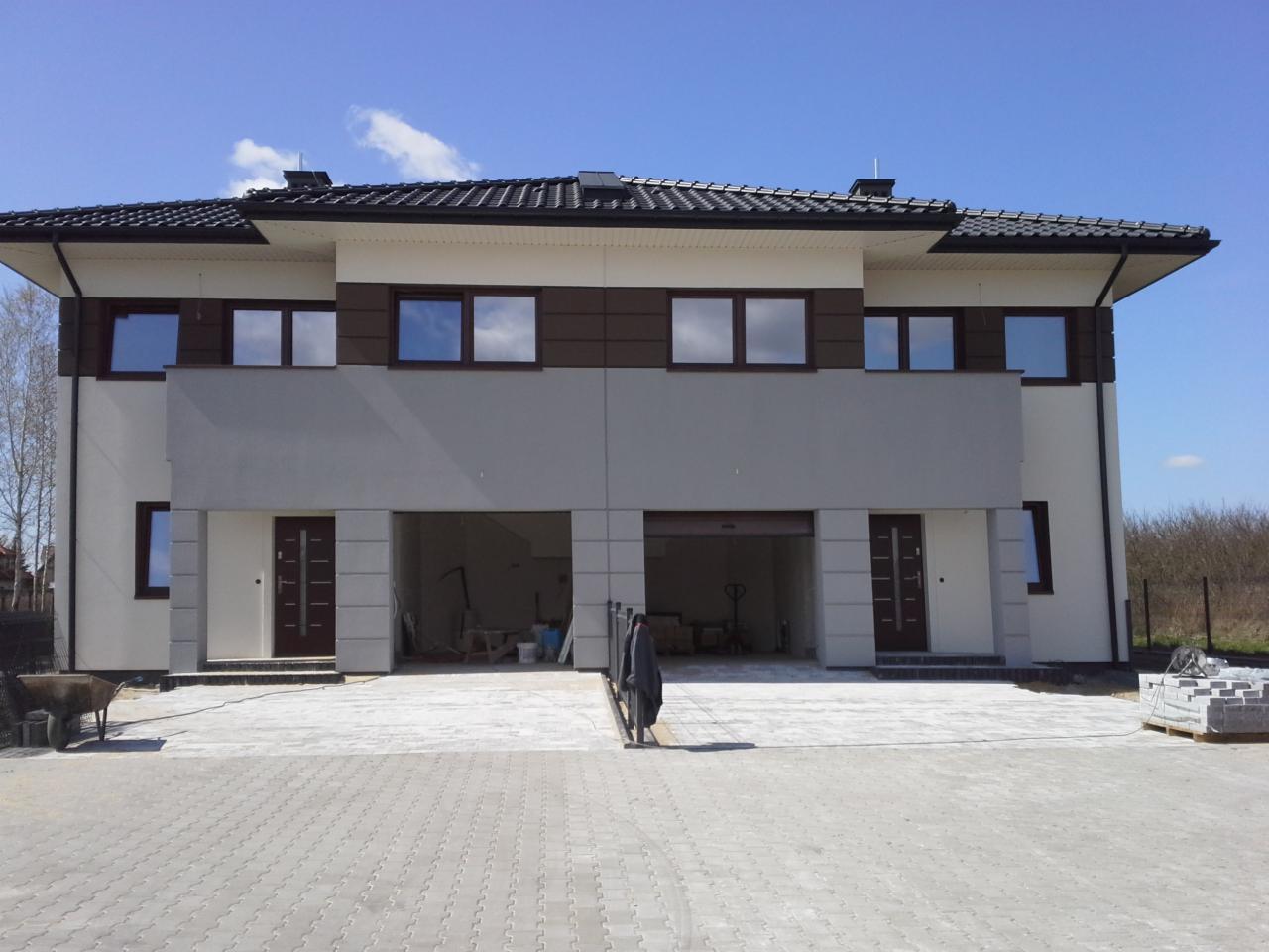 Dwukondygnacyjny dom w zabudowie bliźniaczej z otwartymi garażami i kostką brukową przed budynkiem, widoczne materiały budowlane.