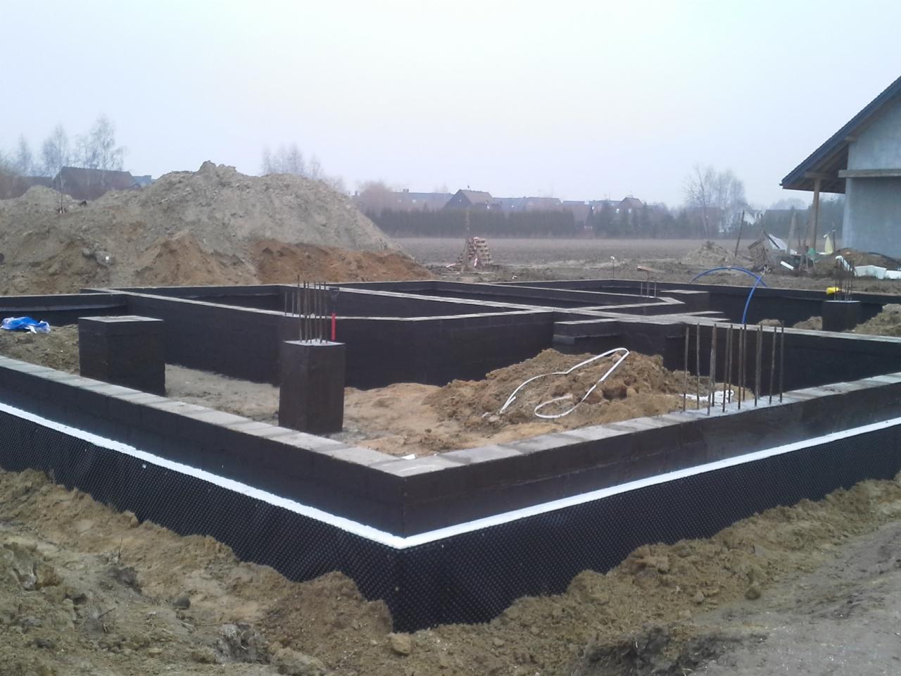 Zabezpieczone fundamenty domu w trakcie budowy, widoczne zbrojenia wystające z betonowych słupków, izolacja pionowa z folii kubełkowej z białym paskiem na górze, w tle hałda ziemi i częściowo...