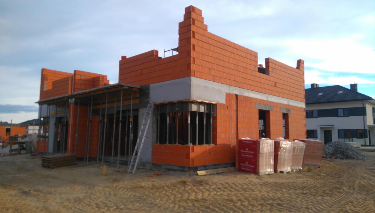 Wznoszona konstrukcja z cegły ceramicznej Porotherm na etapie budowy ścian, widoczne szalunki okienne, drabina oparta o ścianę, palety z cegłami i sąsiedni dom w tle.
