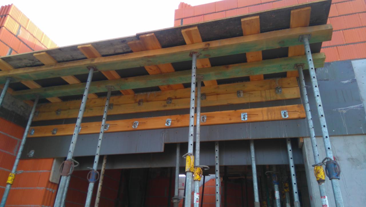 Szare stalowe stemple podtrzymują drewniany szalunek stropu na budowie domu z czerwonej cegły.