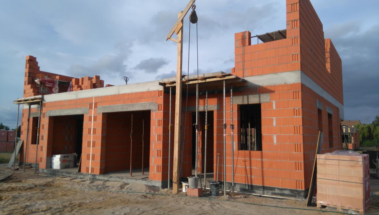 Budowa domu jednorodzinnego z czerwonej cegły, widoczne ściany w stanie surowym, otwory okienne i drzwiowe, drewniany dźwig z linami, podpory stropu, palety z cegłami oraz piasek.