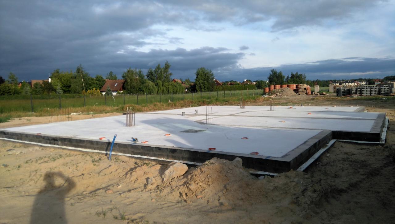 Wylana płyta fundamentowa z wystającymi prętami zbrojeniowymi na działce z widokiem na domy w oddali pod pochmurnym niebem.