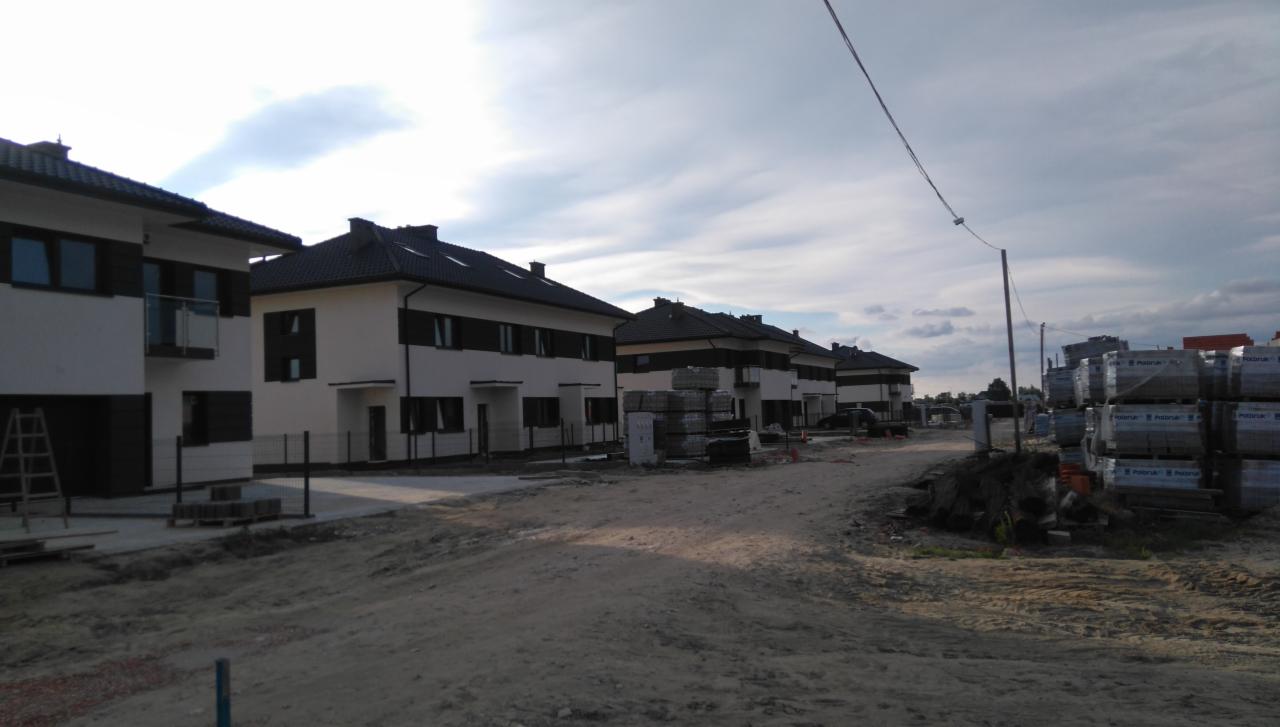 Szeregówka nowych, dwukondygnacyjnych domów z ciemnymi dachami i oknami, w trakcie wykończenia, z widocznymi paletami z materiałami budowlanymi na piaszczystym terenie.