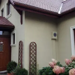 Beżowa elewacja domu jednorodzinnego z brązowymi elementami wykończeniowymi, widoczne okna z białymi ramami, wejście z zadaszeniem i drewnianymi drzwiami oraz ozdobne kratki i krzewy hortensji.