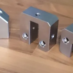 Precyzyjnie wykonane, srebrne, metalowe elementy z otworami, prawdopodobnie frezowane, leżące na drewnianej powierzchni. Różne kształty, dokładne wykończenie.