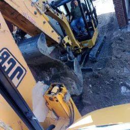 Żółta koparka CAT podczas prac ziemnych w Zielonczynie, widoczny operator w kabinie i fragment wykopu w pobliżu ceglanego budynku.