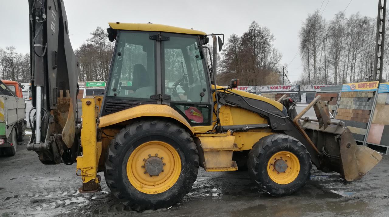 Żółta koparko-ładowarka na placu budowlanym, widok z boku, z widoczną łyżką i ramieniem koparki, w tle kostka brukowa i drzewa.