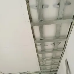 Montaż sufitu podwieszanego z płyt kartonowo-gipsowych na metalowej konstrukcji, widoczne profile i fragment płyty z zaznaczonymi liniami cięcia.