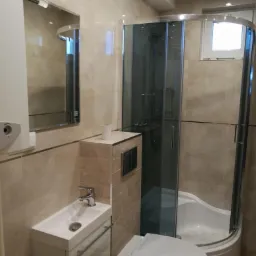 Nowoczesna łazienka z kabiną prysznicową narożną, wiszącą toaletą i umywalką z szafką, wykończona beżowymi płytkami ceramicznymi.