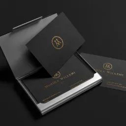 Wizytówki z czarnego papieru ze złotym logo i danymi kontaktowymi, umieszczone w metalowym wizytowniku na ciemnym tle. Minimalistyczny design, elegancka prezentacja.