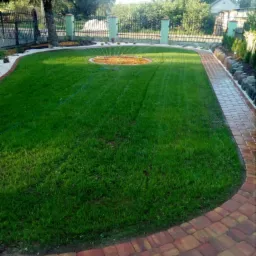 Zadbany trawnik z nawadnianiem w ogrodzie, obrzeża z kostki brukowej, ścieżka z czerwonej kostki, widoczne ogrodzenie i drzewa w tle.