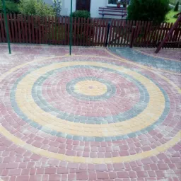 Kolorowa kostka brukowa ułożona w okrągły wzór na podwórku przed domem z drewnianym płotem.