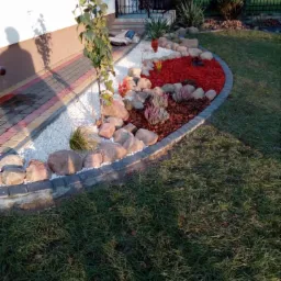 Aranżacja ogrodu z obrzeżem z kostki, białym i czerwonym grysem, kamieniami i młodym drzewkiem liściastym przy elewacji domu.