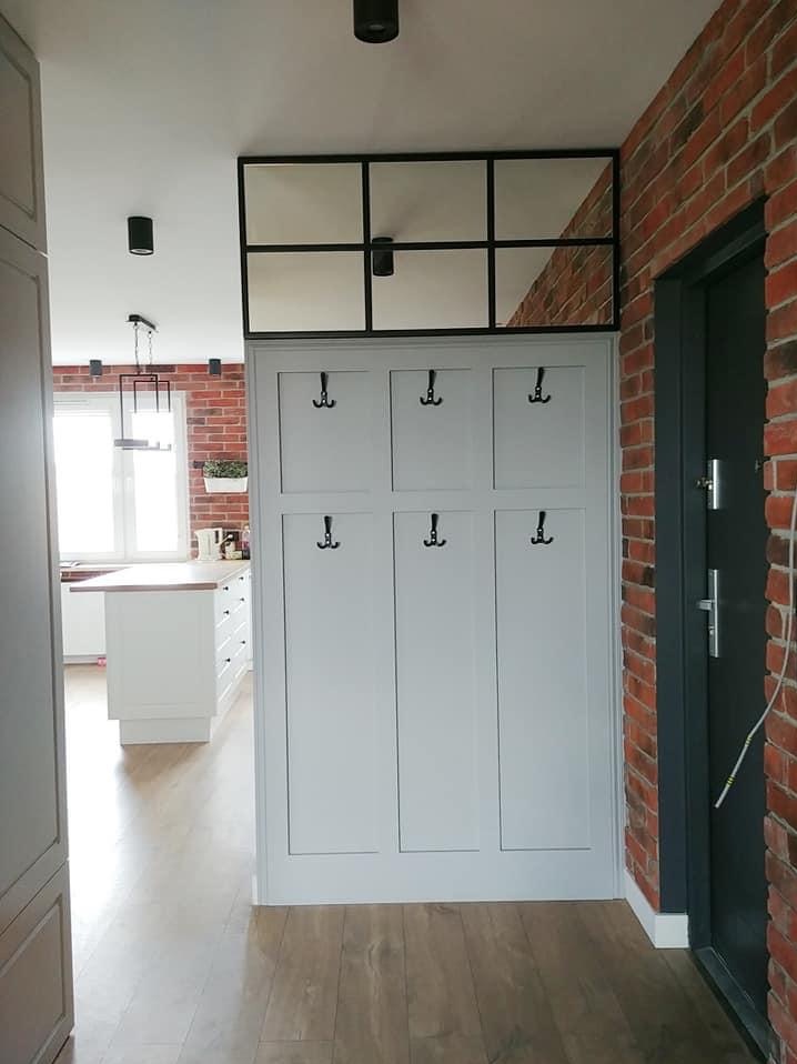 Szara zabudowa z wieszakami w przedpokoju, widok na kuchnię w tle, ceglana ściana z boku, Gdańsk.