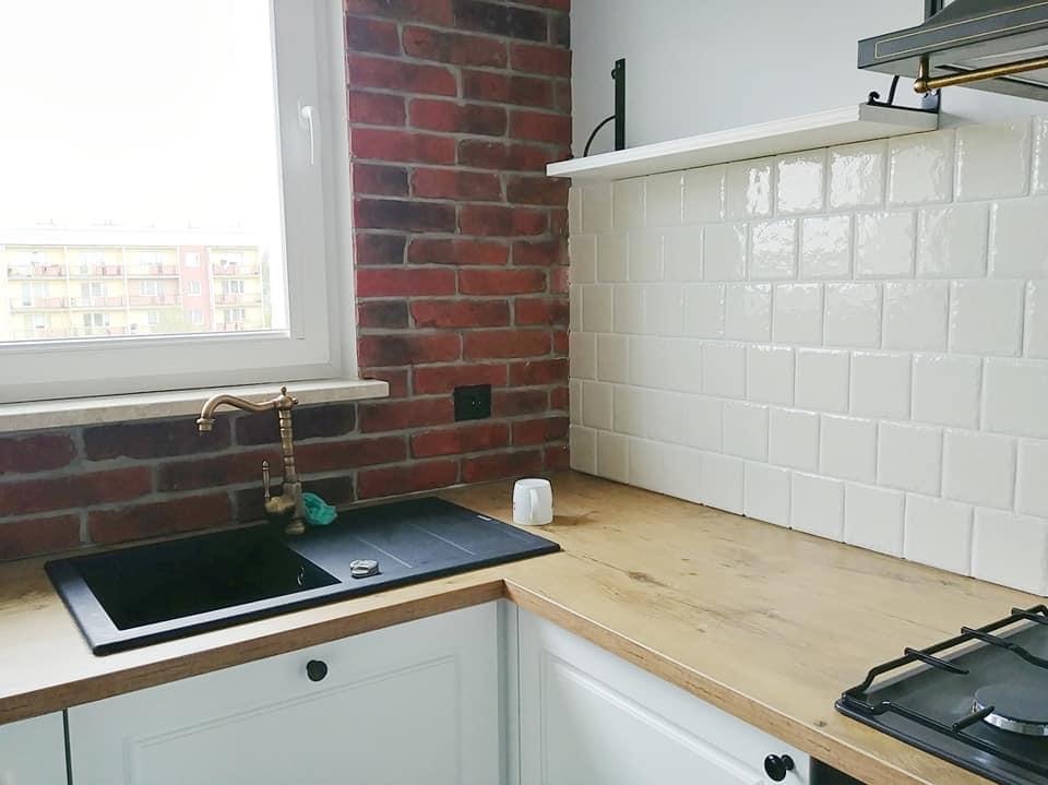 Kuchenny blat z drewnianym wykończeniem, czarny zlew z mosiężną baterią, białe szafki z czarnymi uchwytami, ściana z białych kwadratowych płytek i fragment ściany z czerwonej cegły.