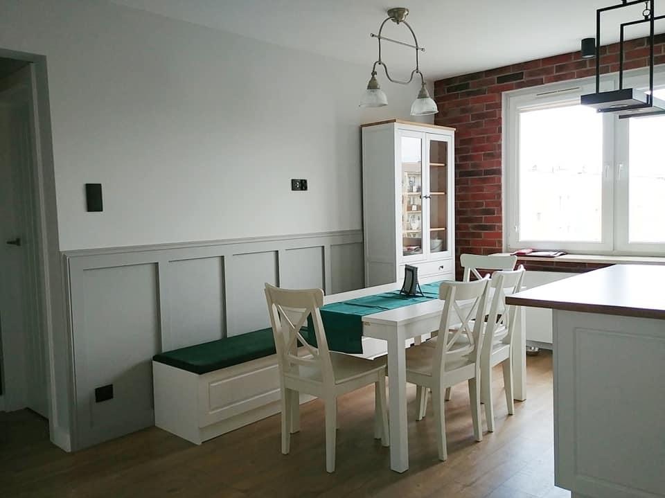 Jasne wnętrze jadalni z białymi meblami: stół z krzesłami, szafka przeszklona, siedzisko w zabudowie z zieloną poduszką. Ściana z cegły przy oknie.