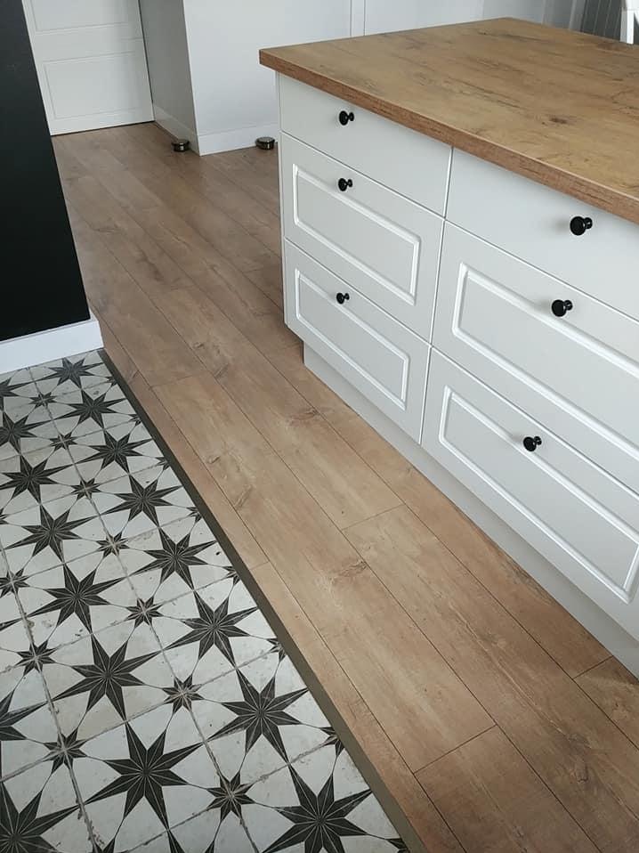 Kontrastowe połączenie podłóg: wzorzyste płytki w gwiazdy i panele imitujące drewno, obok białej szafki kuchennej z drewnianym blatem i czarnymi gałkami.