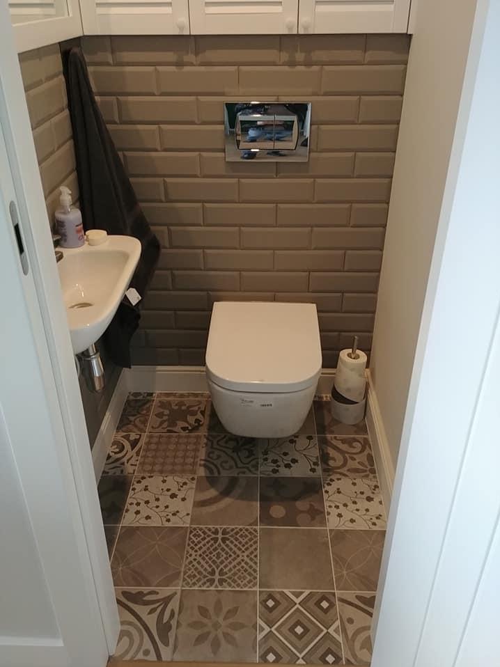 Mała toaleta z mozaikową podłogą, brązowymi płytkami na ścianie i podwieszaną miską WC. W rogu widoczny stojak na papier toaletowy. Nad toaletą panel sterujący spłuczką.