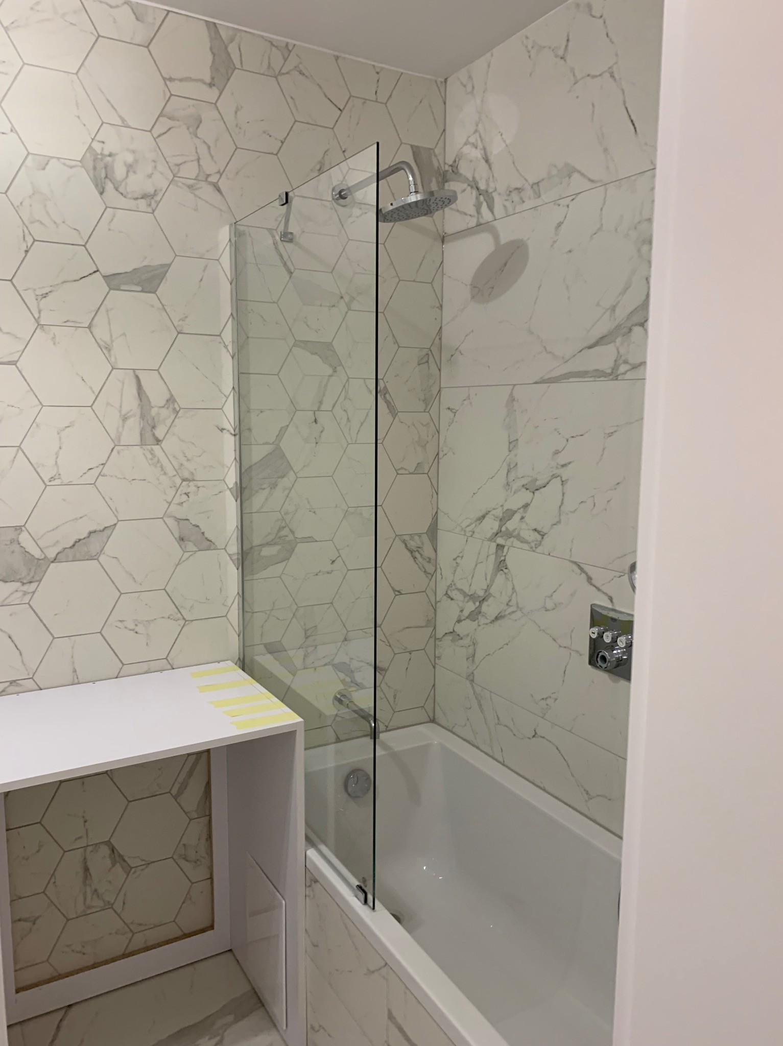 Łazienka z wanną obudowaną płytkami imitującymi marmur, przeszklony parawan nawannowy, bateria prysznicowa z deszczownicą oraz biała szafka z żółtą taśmą malarską na blacie.