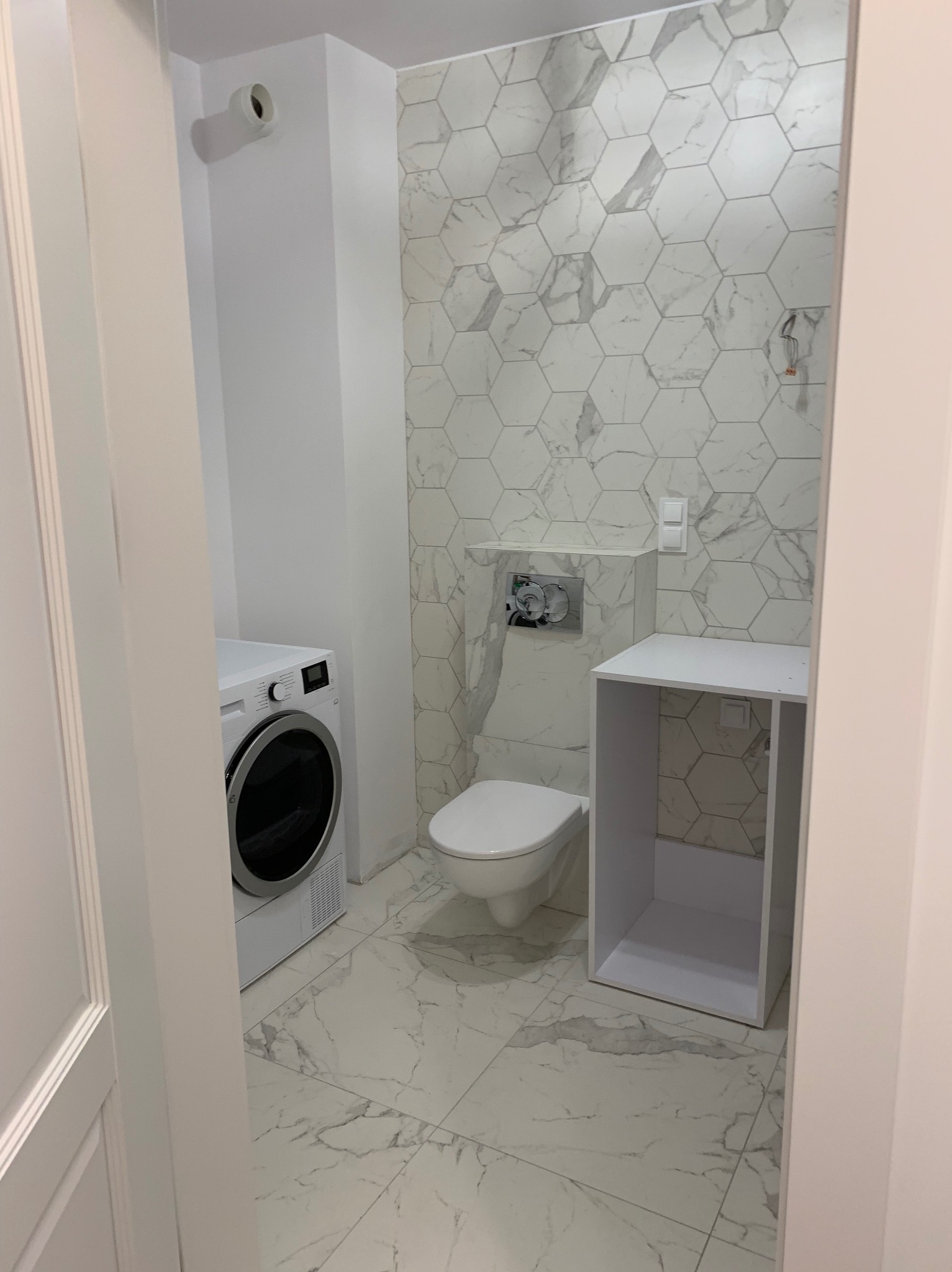 Wykończona łazienka w bieli i szarości z heksagonalnymi płytkami marmurowymi na ścianie, widok na toaletę, pralkę i szafkę bez drzwi.