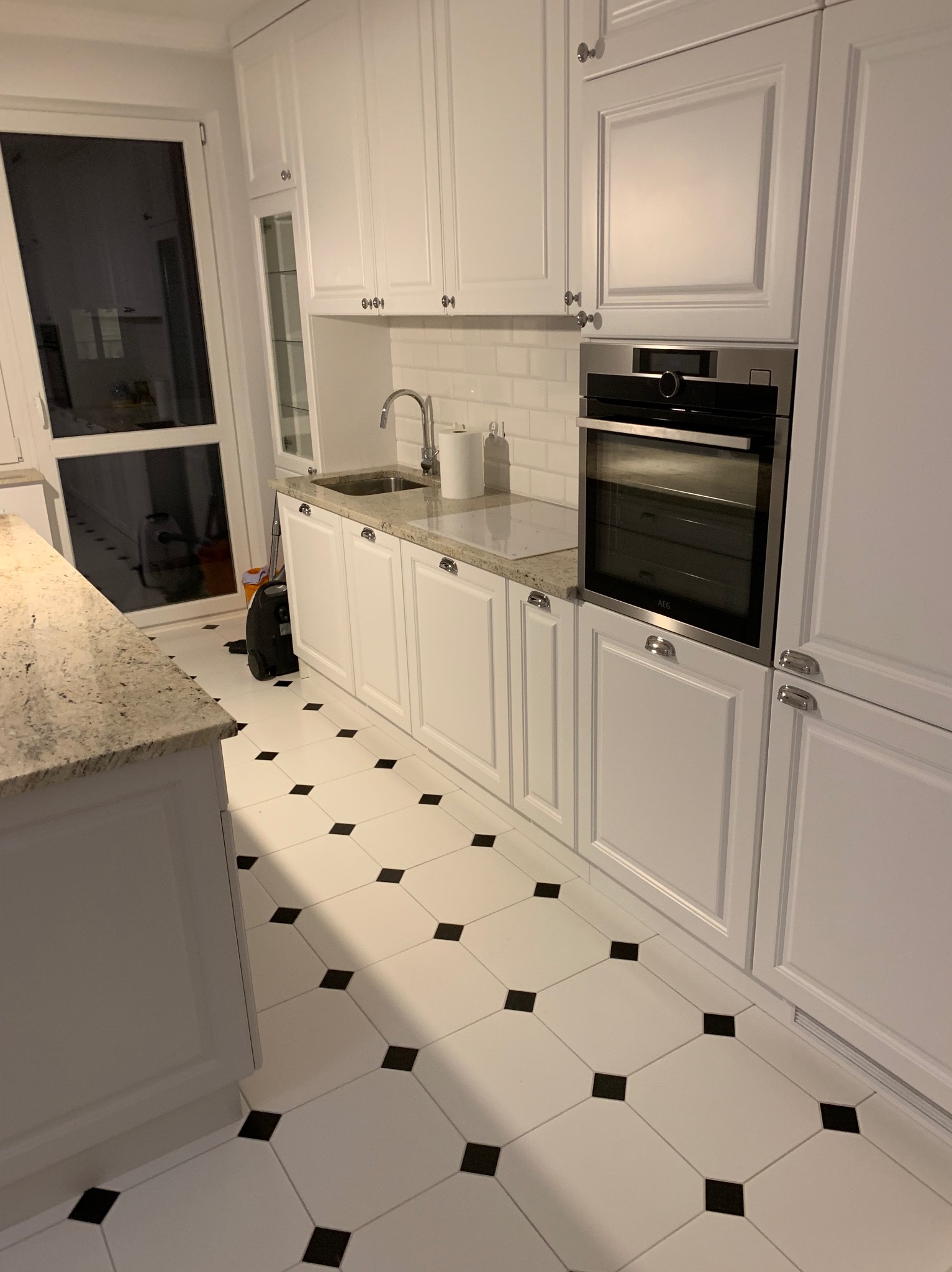 Biała zabudowa kuchenna z piekarnikiem AEG, blatem z jasnego granitu i biało-czarną podłogą w romby, widok na zlew i okno.