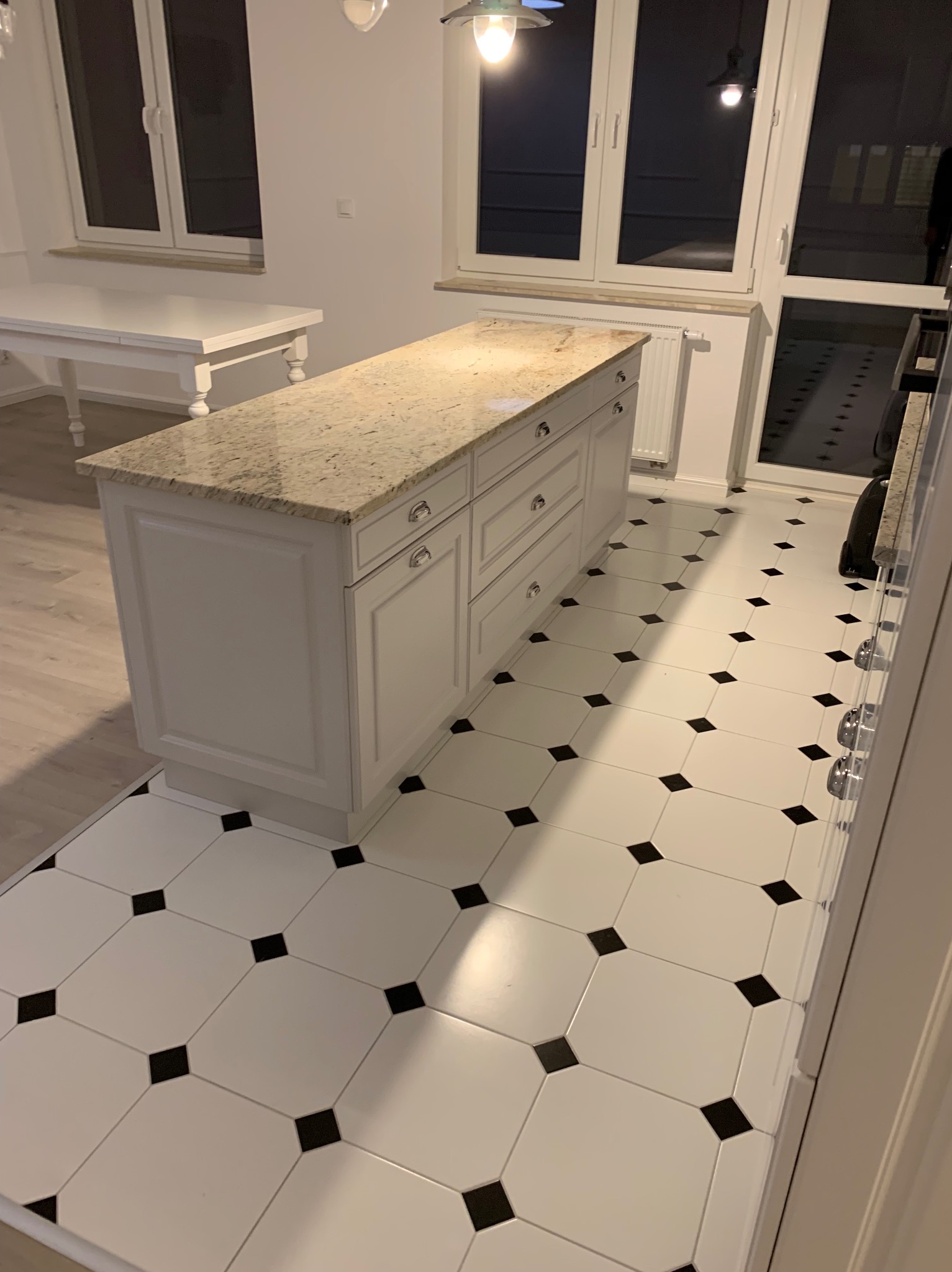 Biała wyspa kuchenna z blatem z jasnego granitu, klasyczne frezowane fronty szuflad i szafek, biała podłoga w czarno-białą szachownicę, widok na okno i stół w tle.