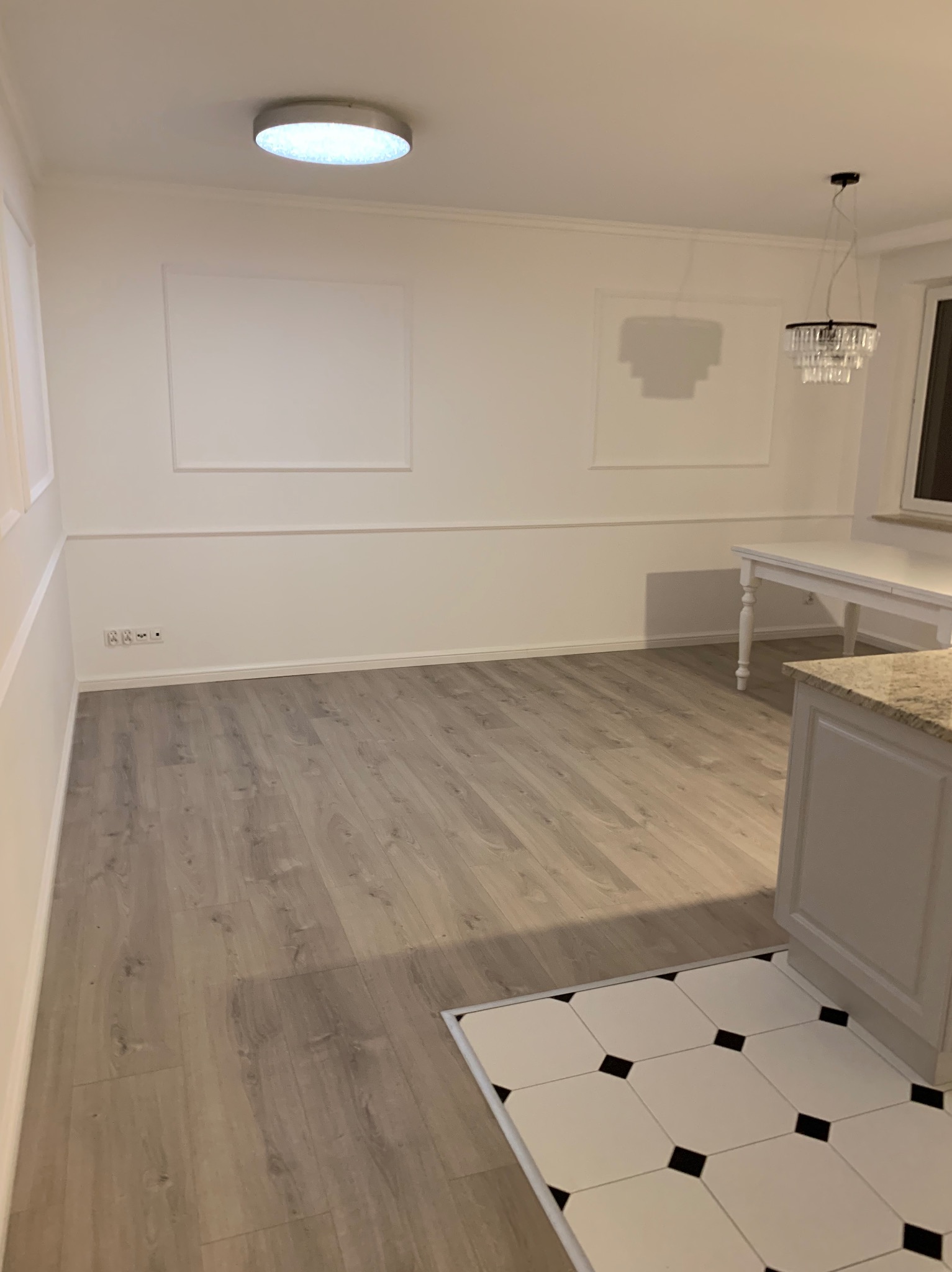 Jasne wnętrze po remoncie: panele podłogowe imitujące drewno, białe ściany z dekoracyjnymi panelami, nowoczesny żyrandol i fragment zabudowy kuchennej z blatem imitującym kamień.