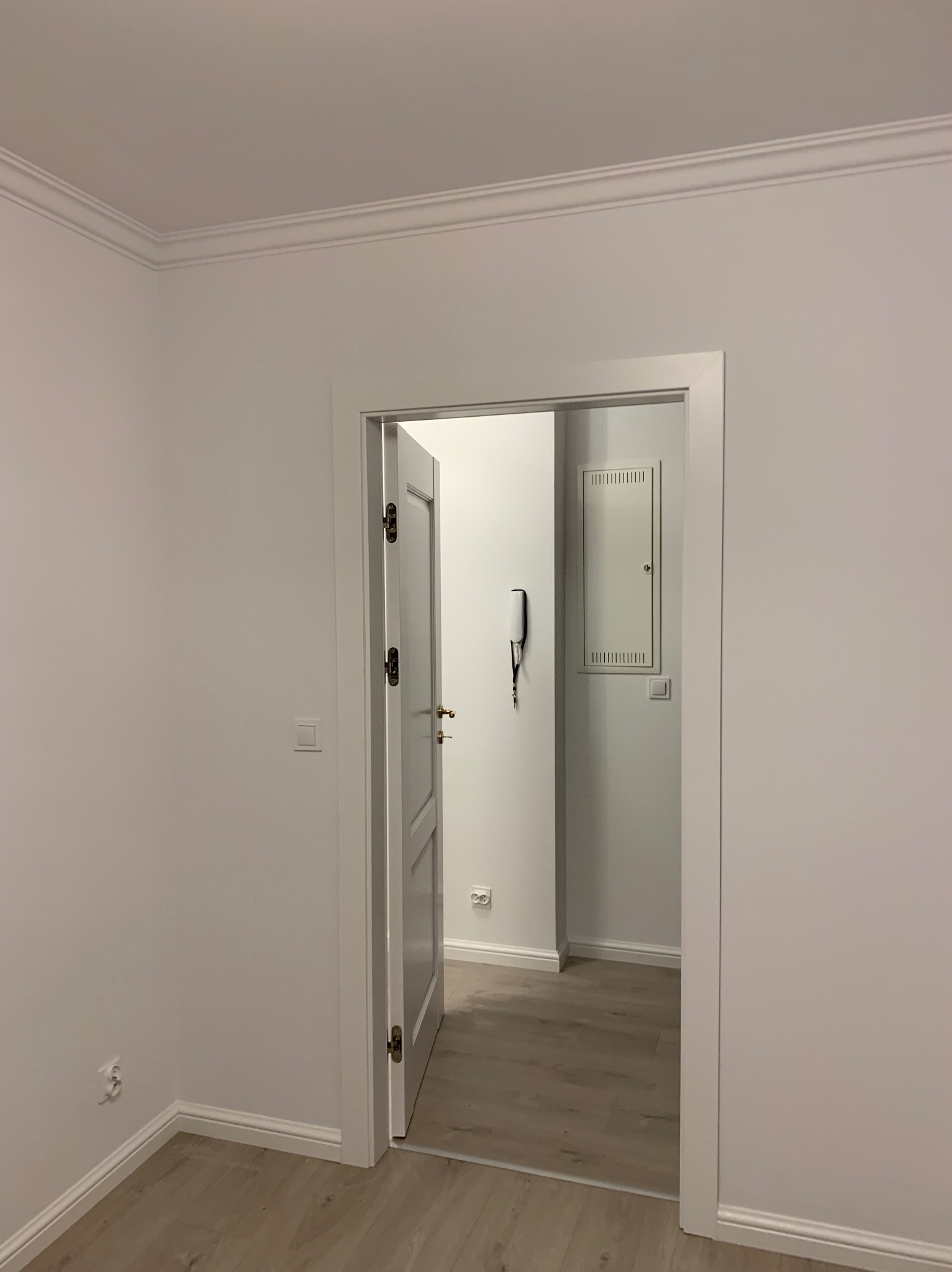 Białe, odświeżone wnętrze z otwartymi drzwiami, widoczne listwy przypodłogowe i sufitowe, jasna podłoga imitująca drewno, skrzynka elektryczna i domofon w głębi pomieszczenia.