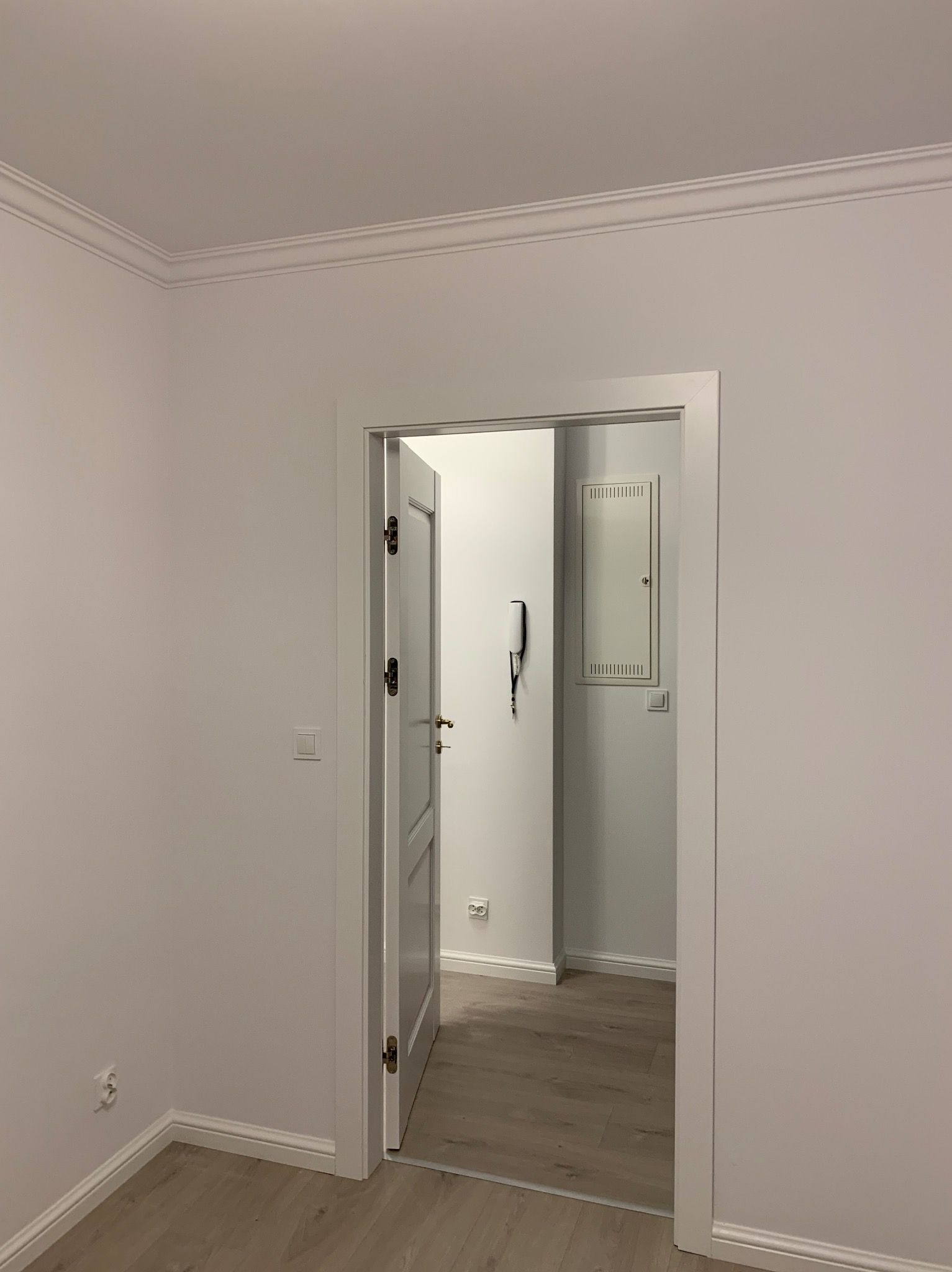 Wnętrze po odświeżeniu: białe ściany z dekoracyjną listwą przysufitową, otwarta biała rama drzwiowa, widoczny fragment podłogi z paneli w odcieniach beżu, w tle skrzynka elektryczna i domofon.