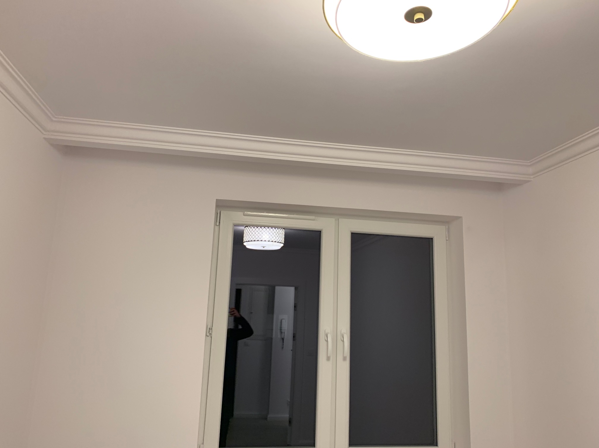 Białe listwy dekoracyjne sufitowe zamontowane w rogu pokoju, widoczny fragment sufitu z okrągłą lampą, okno z widocznym odbiciem osoby.