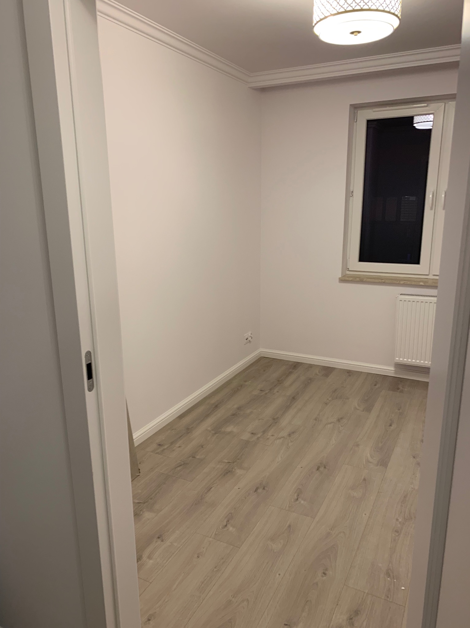 Pusty pokój po remoncie z panelami podłogowymi w odcieniach beżu, białymi ścianami z dekoracyjną listwą przysufitową i grzejnikiem pod oknem.