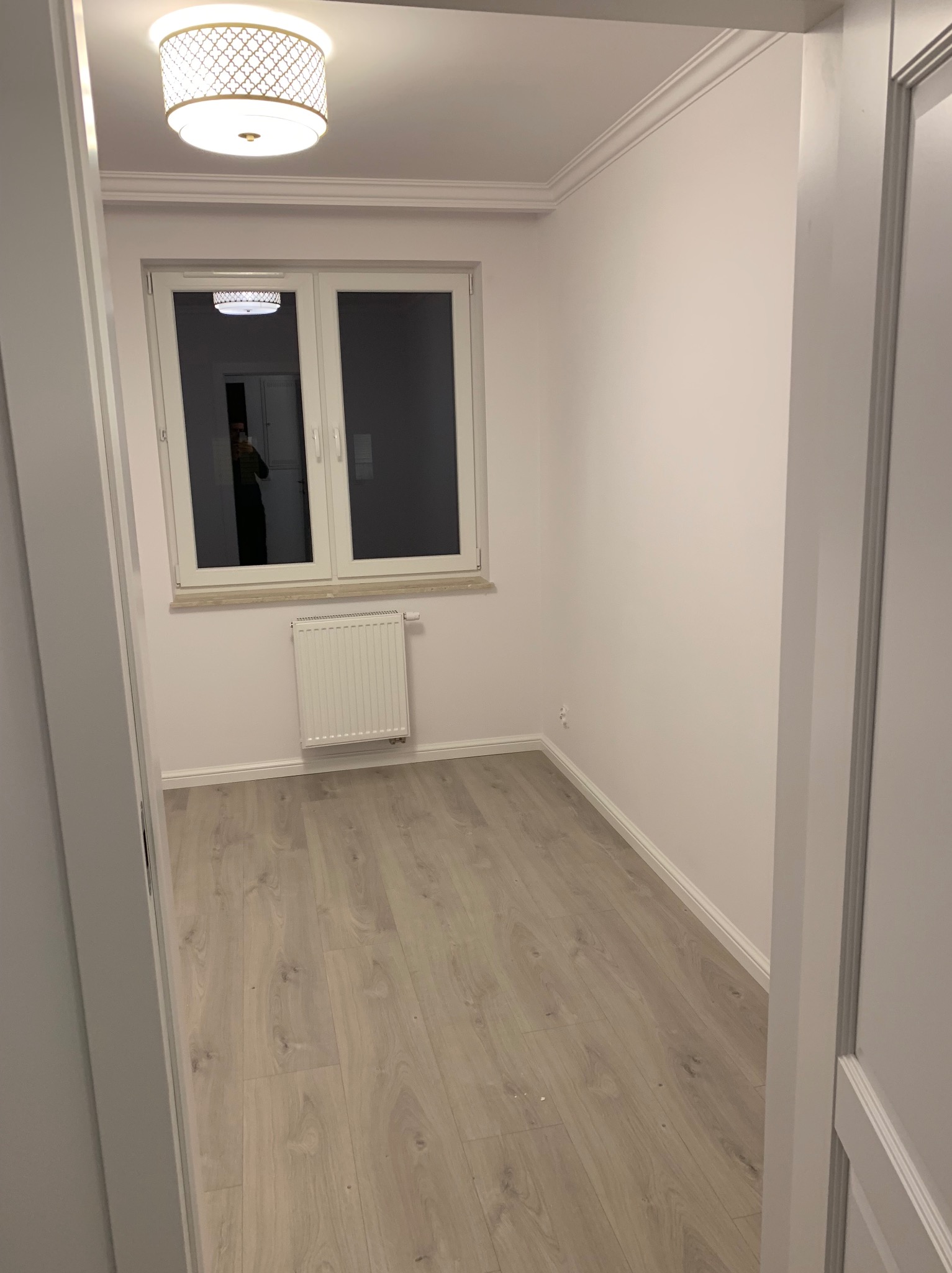 Pusty pokój z jasnymi ścianami, panelami podłogowymi i grzejnikiem pod oknem, widok zza otwartych drzwi, na suficie ozdobna lampa.