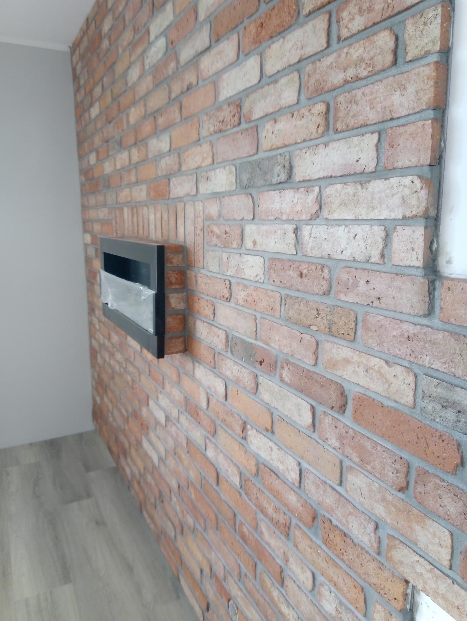 Ściana z cegły klinkierowej w odcieniach brązu i beżu z wnęką na biokominek, szara fuga, widoczne fragmenty szarej ściany i podłogi z paneli.