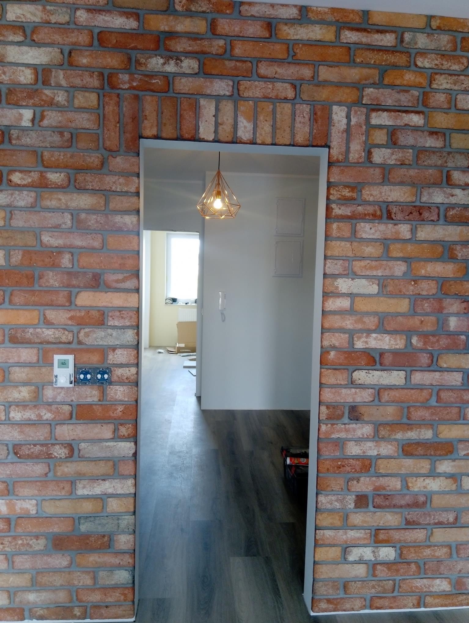 Ściana z cegły klinkierowej wokół wejścia do pokoju z widocznym fragmentem podłogi, lampą wiszącą i elementami instalacji elektrycznej.